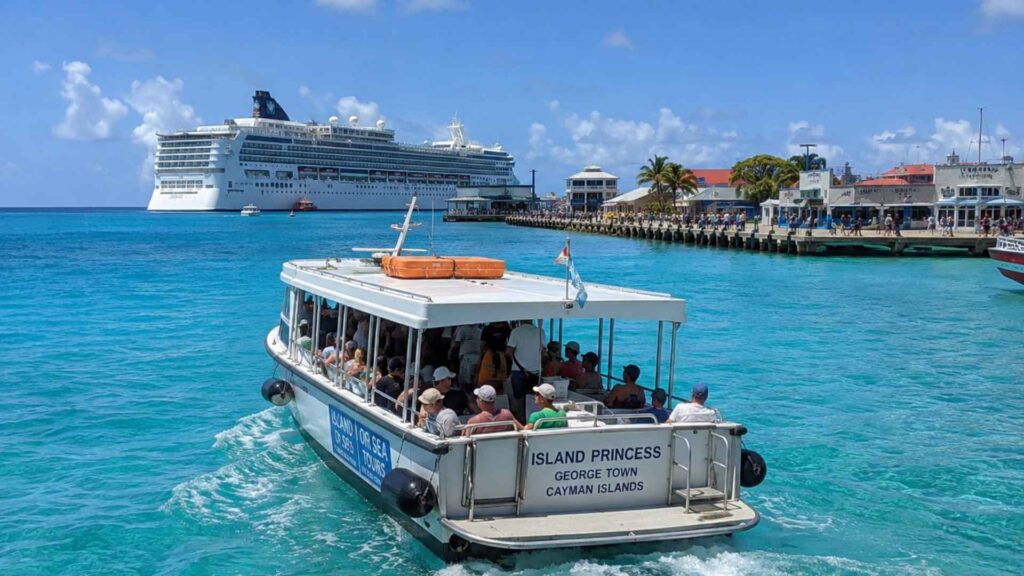 Cruise tender carrying passengers across turquoise water toward George Town, Grand Cayman, with a cruise ship anchored offshore.