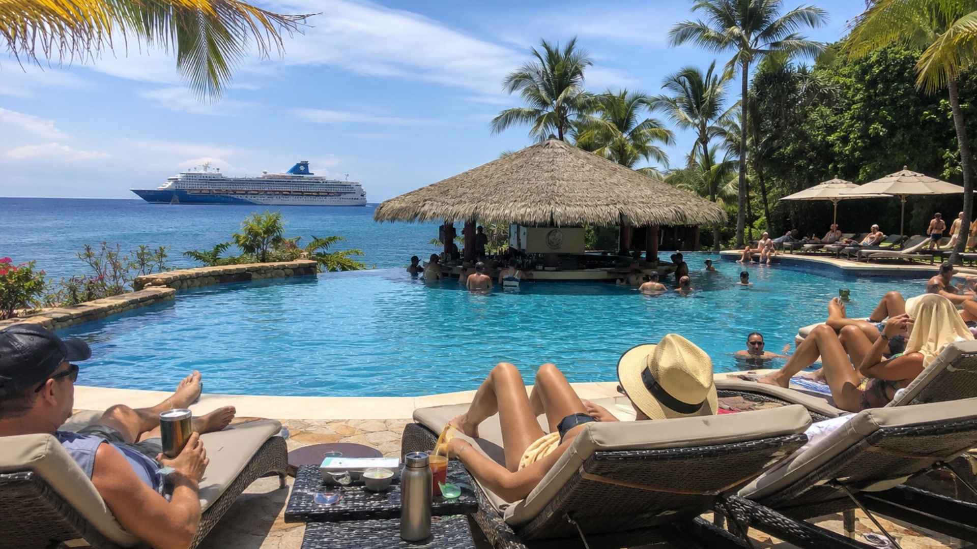 Guests relaxing by a tropical infinity pool with a swim-up bar and an anchored cruise ship visible offshore.