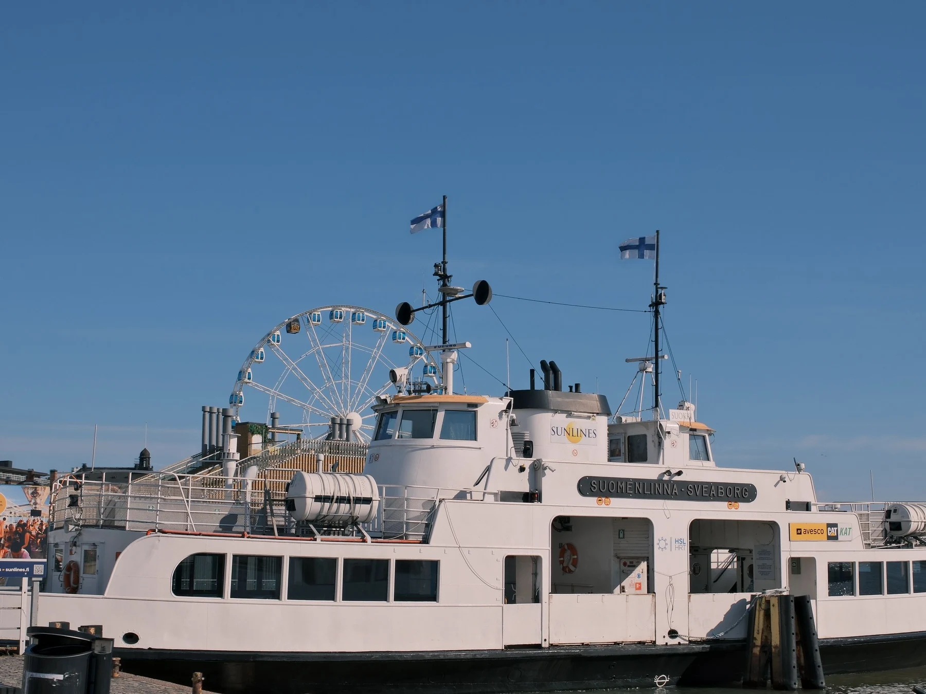 Passenger ferry docked at Helsinki harbor with the SkyWheel and Finnish flags behind it.