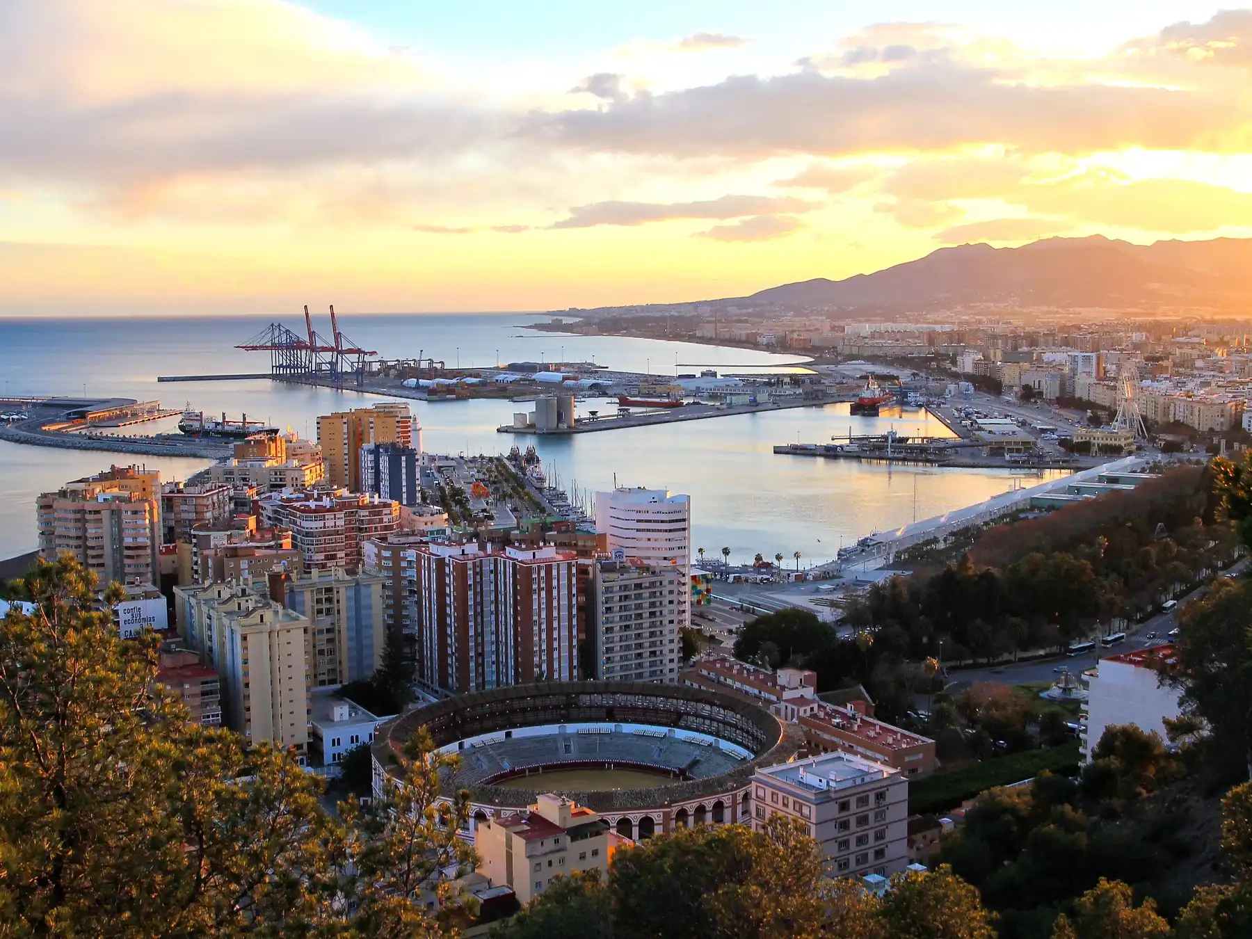 Aerial view of Málaga cruise port, the city waterfront, bullring, and mountains at sunset.