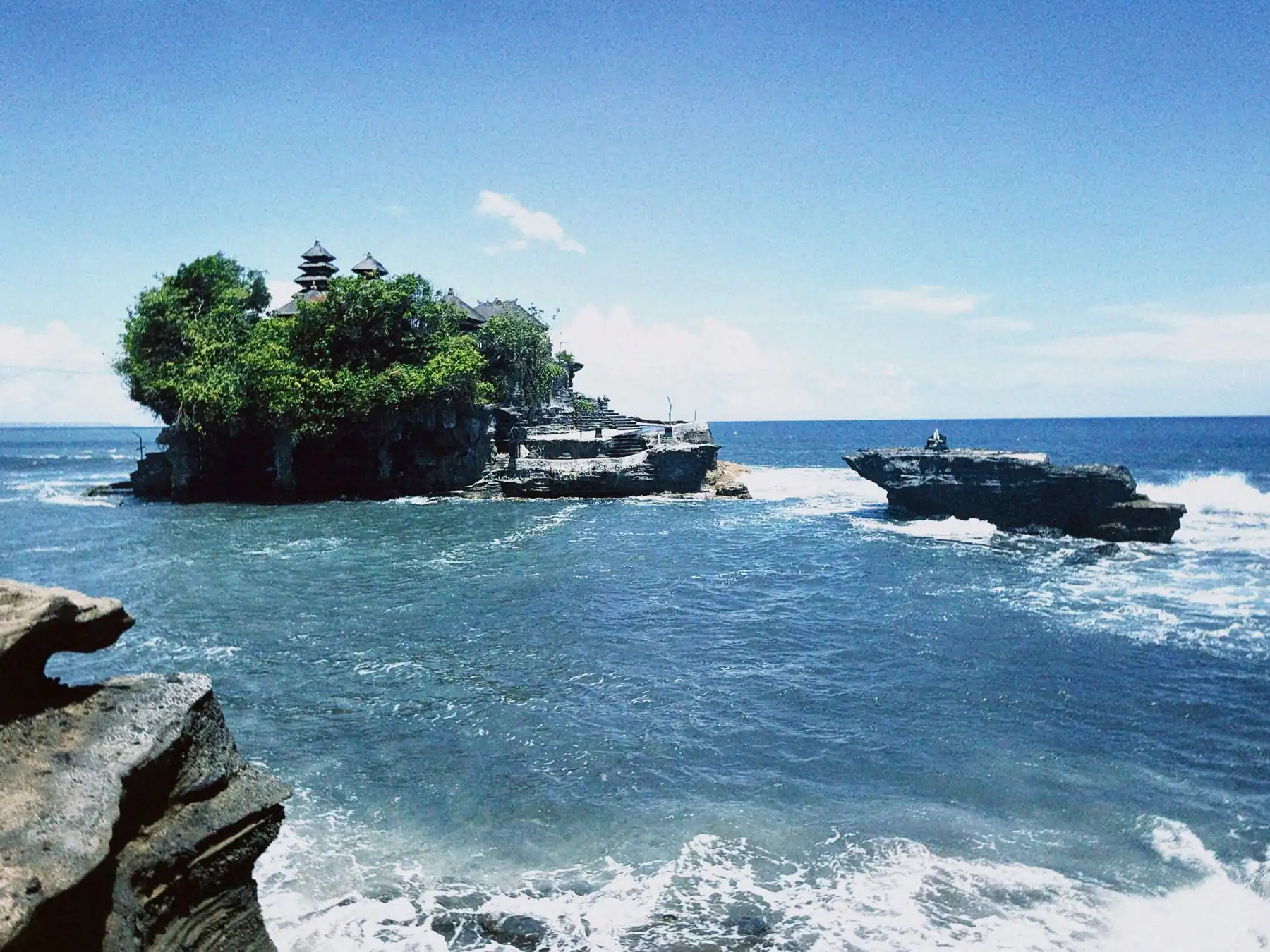 Tanah Lot Temple on Bali's rocky coastline beneath a blue sky.