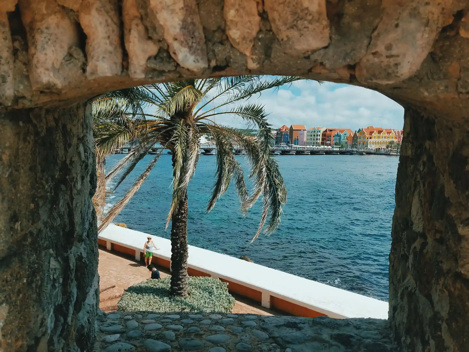Palm trees and colorful Willemstad waterfront buildings seen through a stone arch.