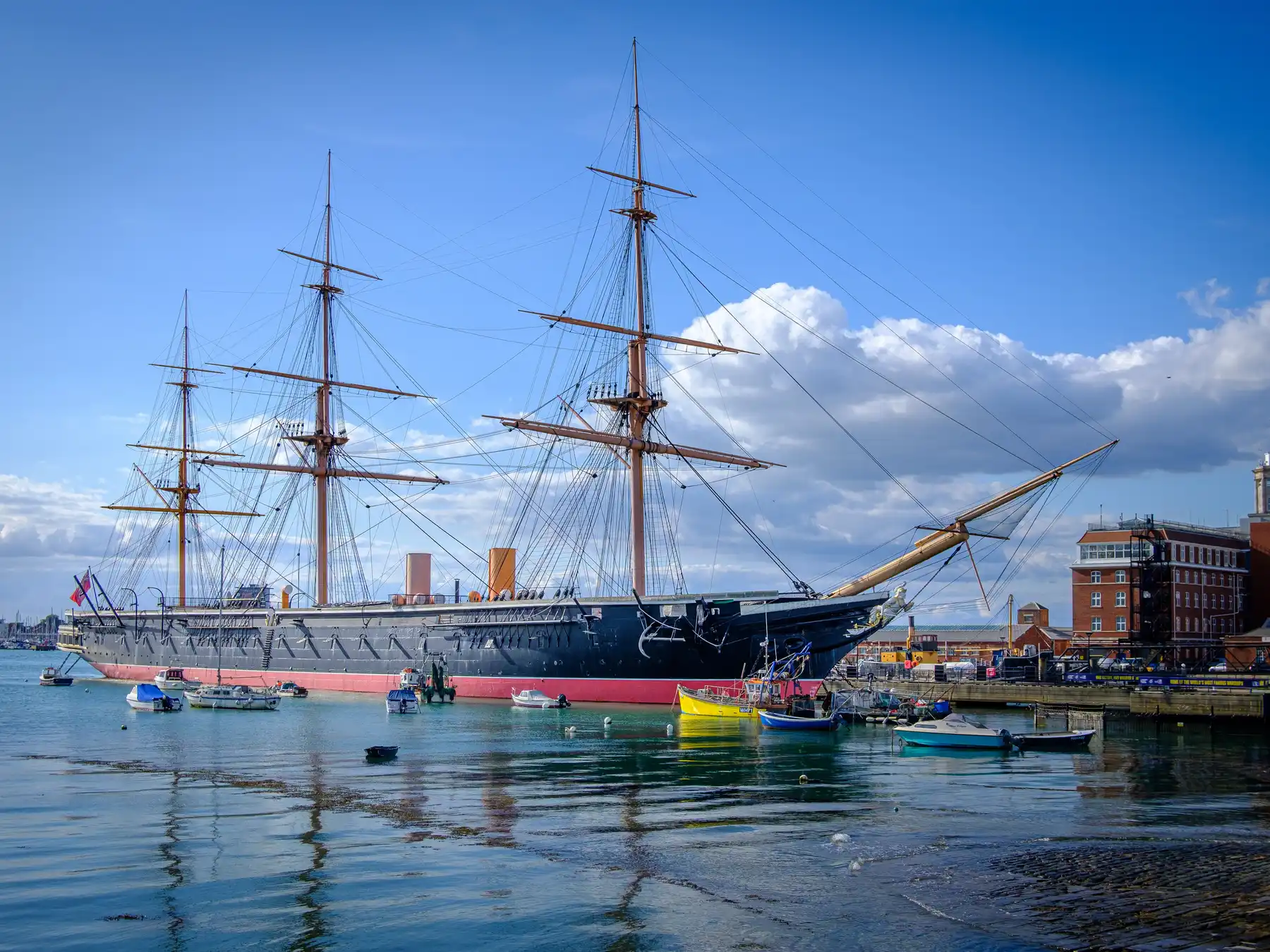 HMS Warrior moored at Portsmouth Historic Dockyard with boats and harbor reflections.