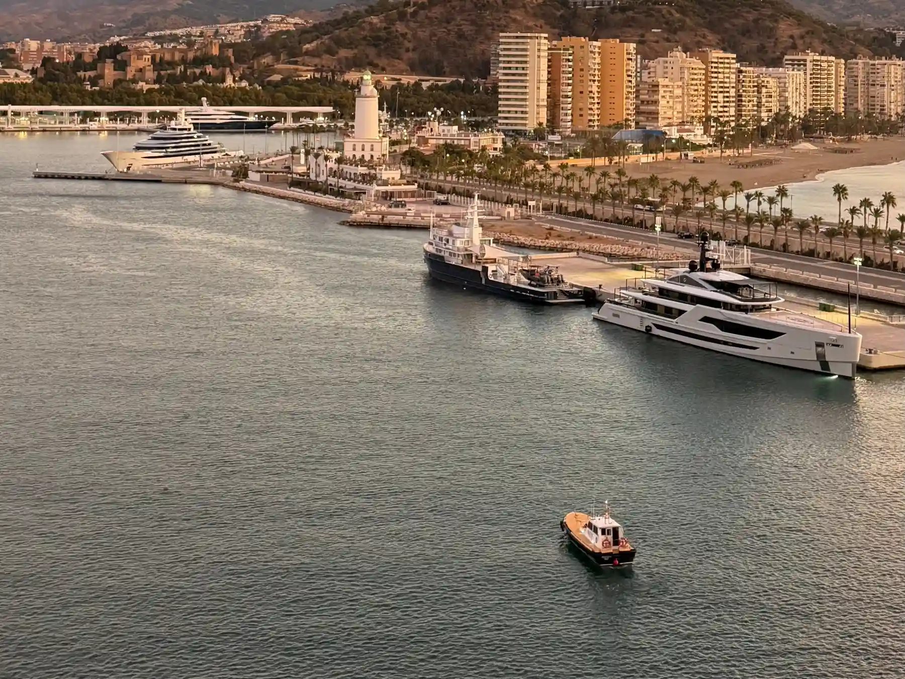 Yachts in Málaga harbor with La Farola lighthouse and the waterfront at sunset.