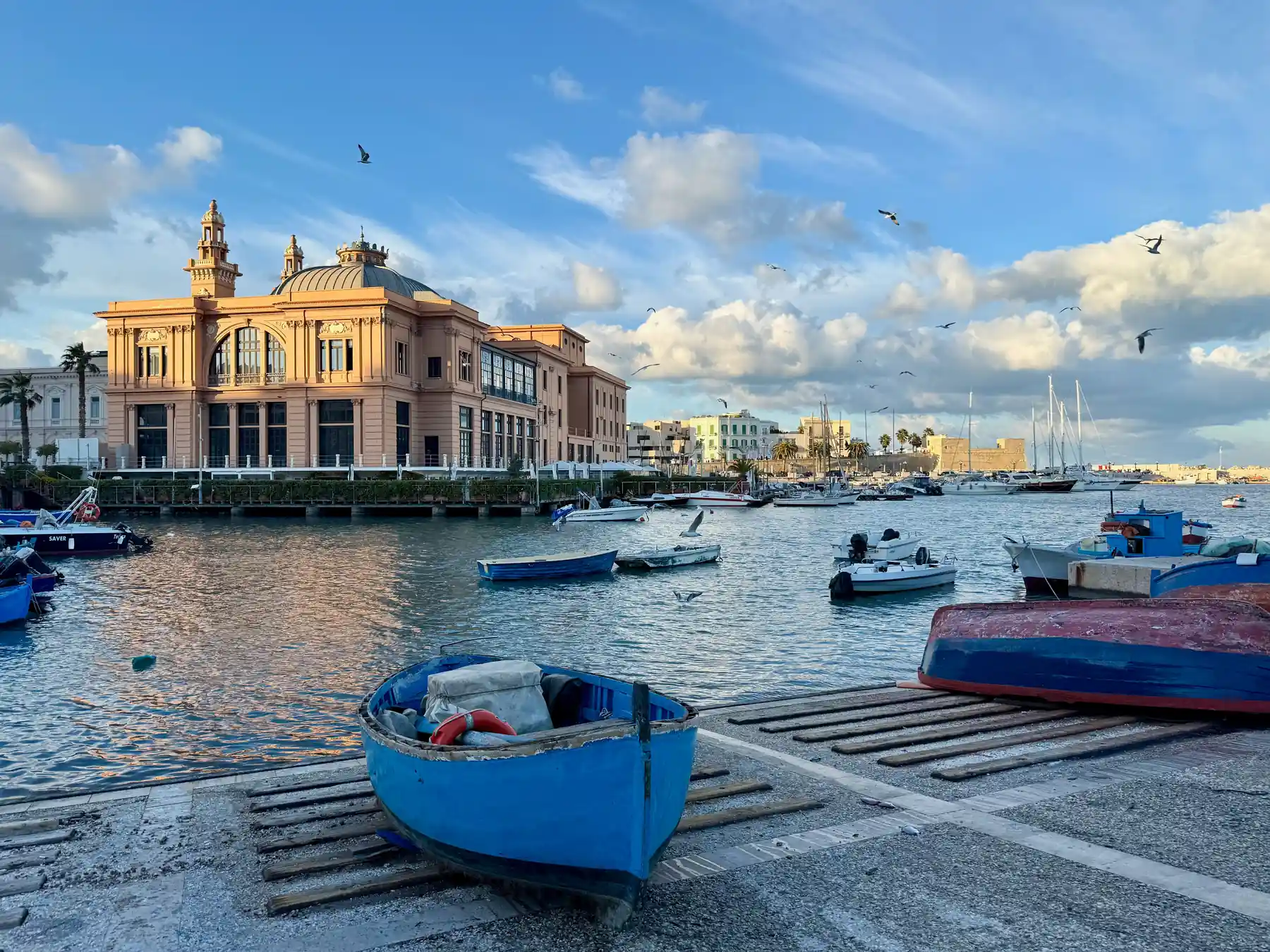 Boats in Bari Harbor beside a historic waterfront building under blue skies.
