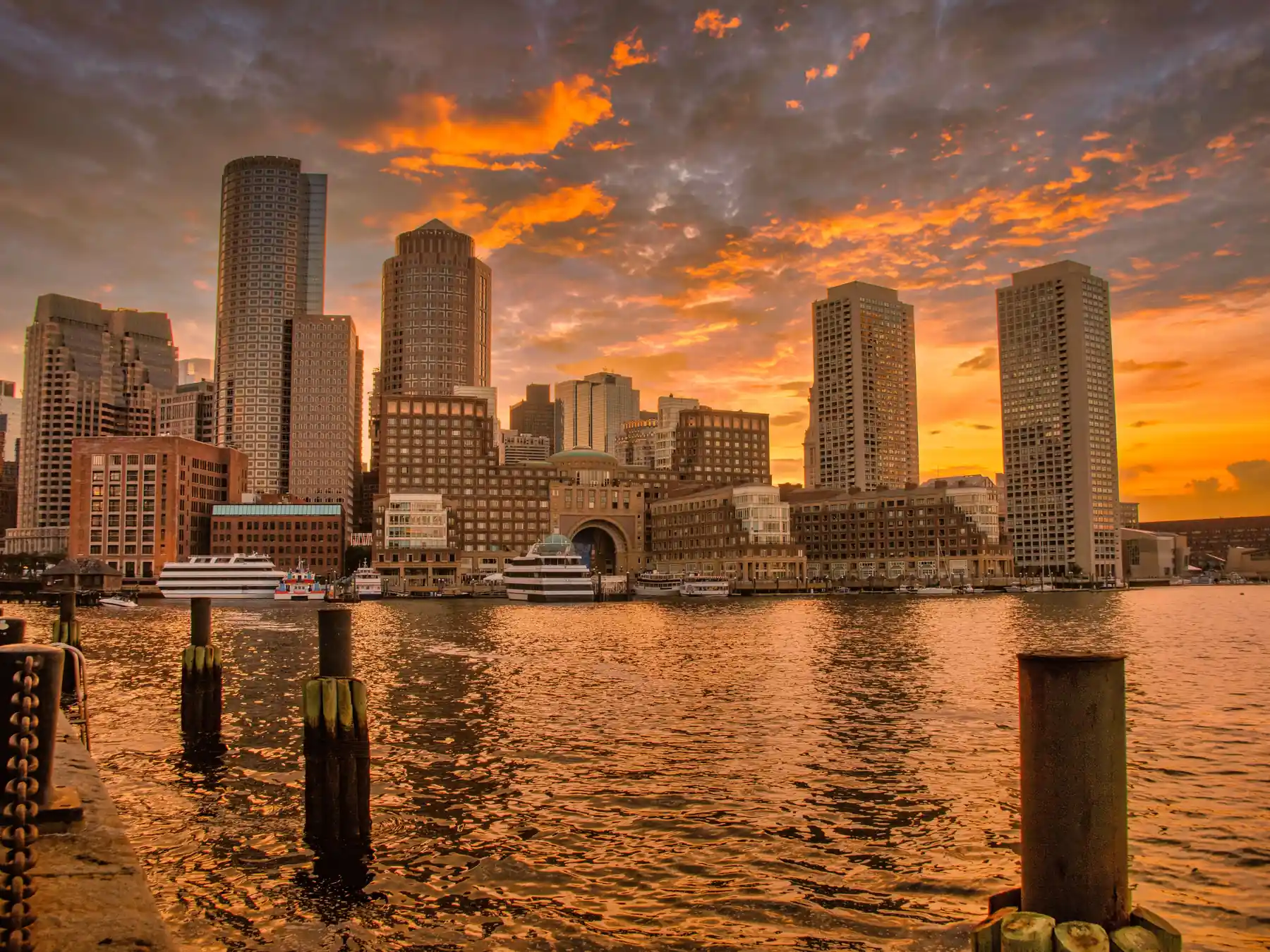 Boston waterfront skyscrapers illuminated by sunset over the harbor.