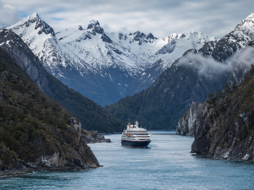Small expedition cruise ship sailing a narrow channel in the Chilean fjords beneath snowy peaks