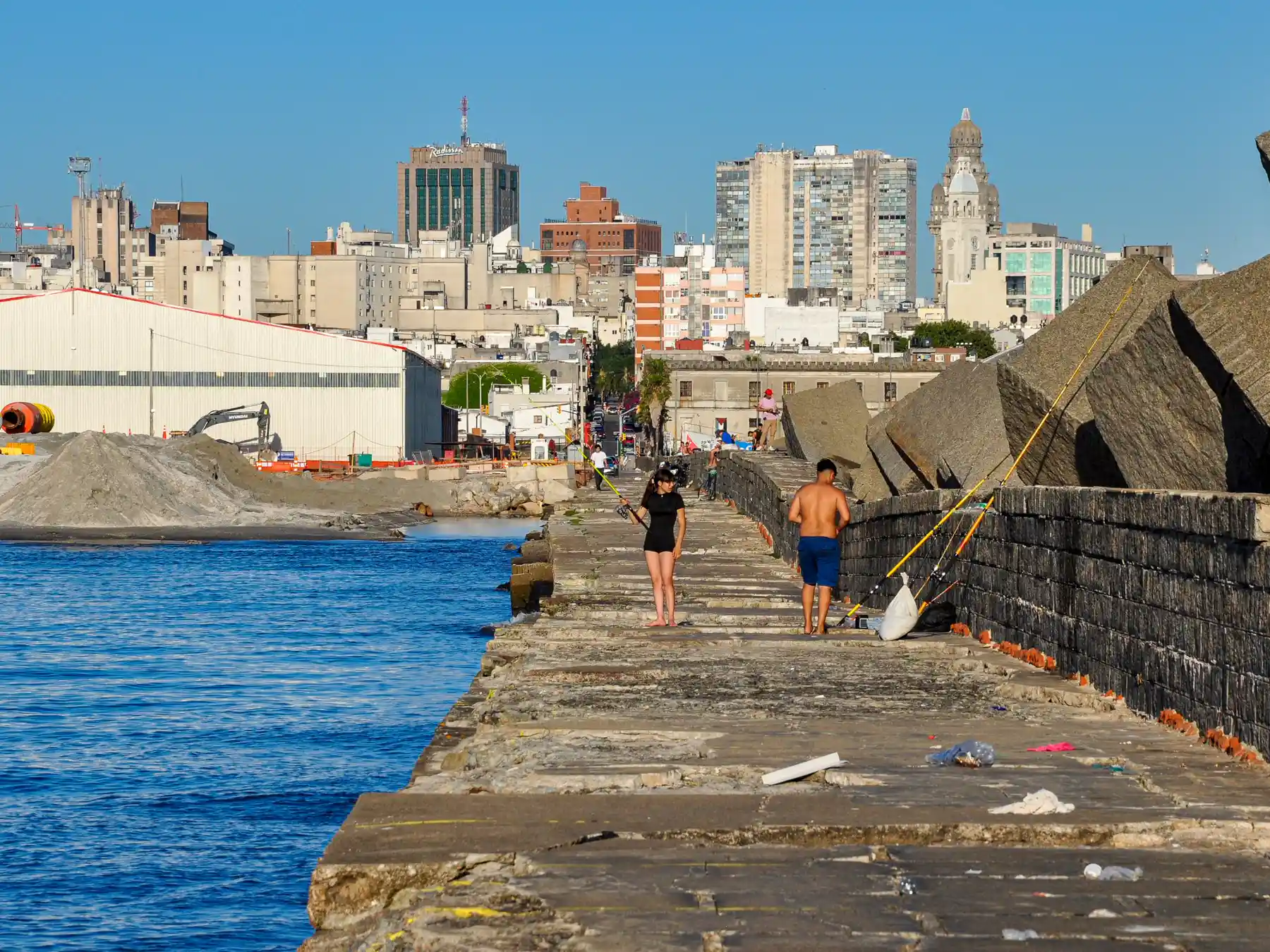 People walking and fishing along a coastal pier with Montevideo's skyline beyond.
