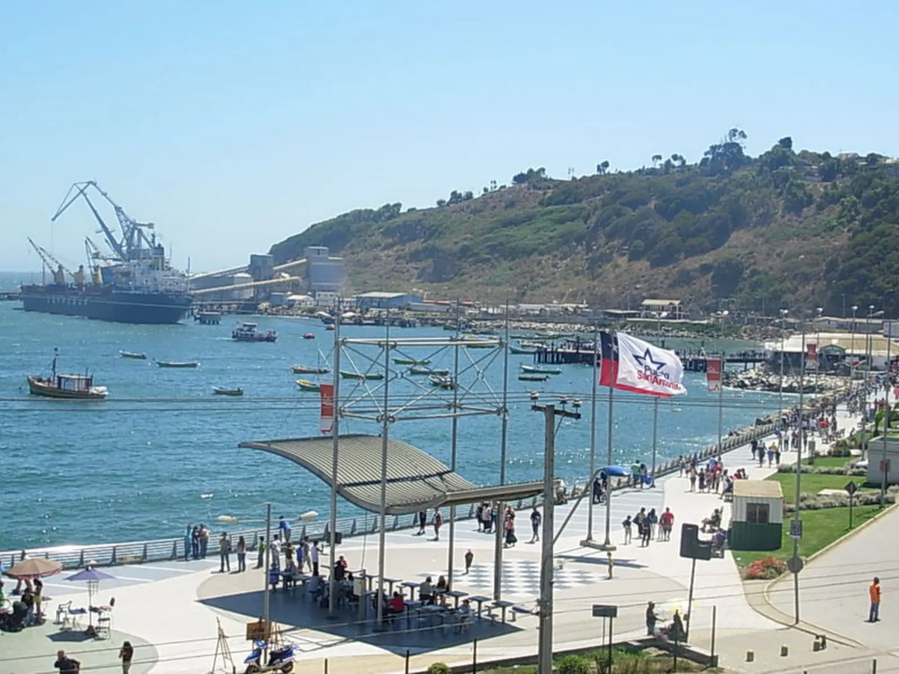 Visitors on San Antonio's waterfront promenade with fishing boats and port cranes behind.
