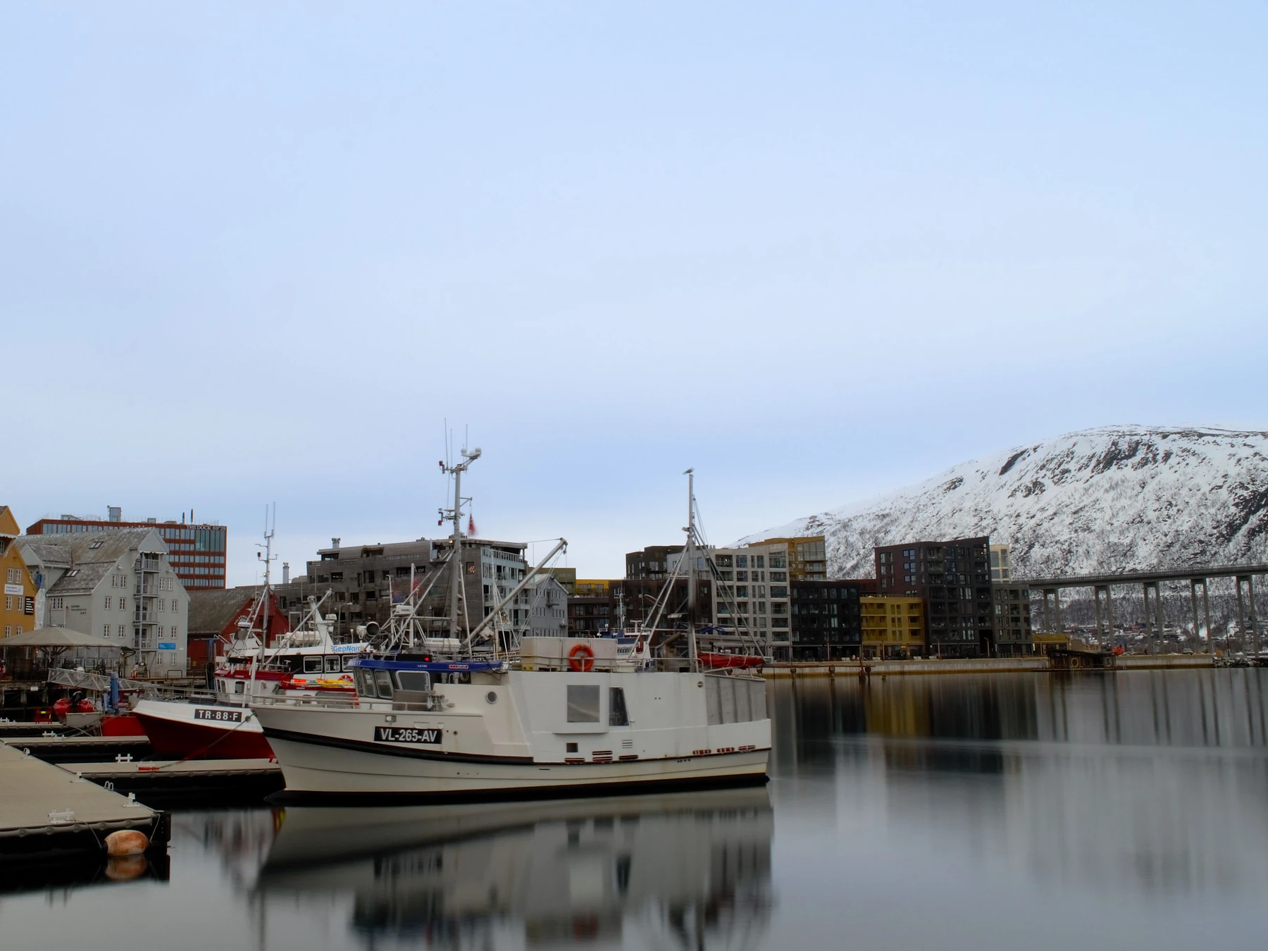 Boats and colorful waterfront buildings in Tromsø harbor beneath snowy mountains.