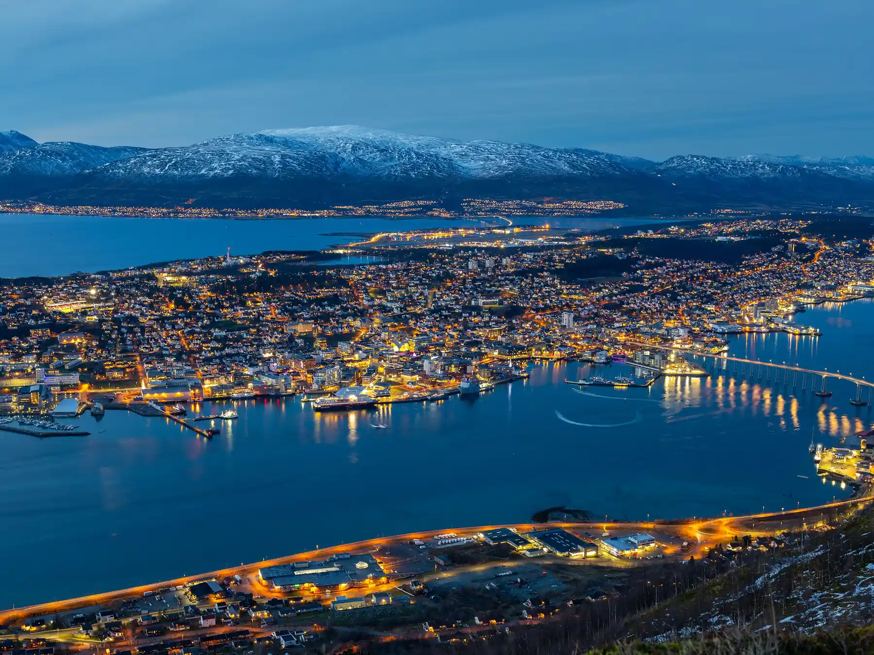 Tromsø's illuminated waterfront, harbor, and mountains reflected at night.