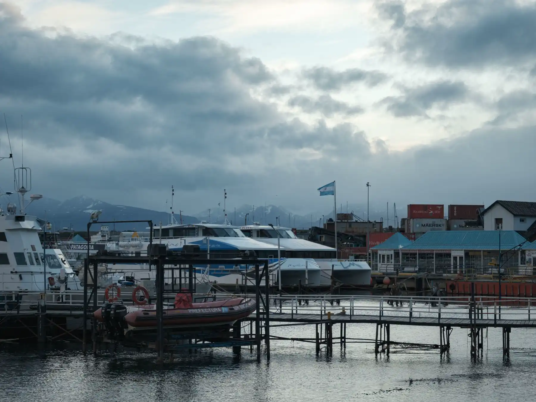 Boats moored at Ushuaia harbor with port buildings and mountains under cloudy skies.