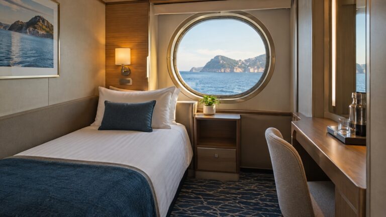Empty single cabin on a cruise ship with one bed and a sea-facing porthole.