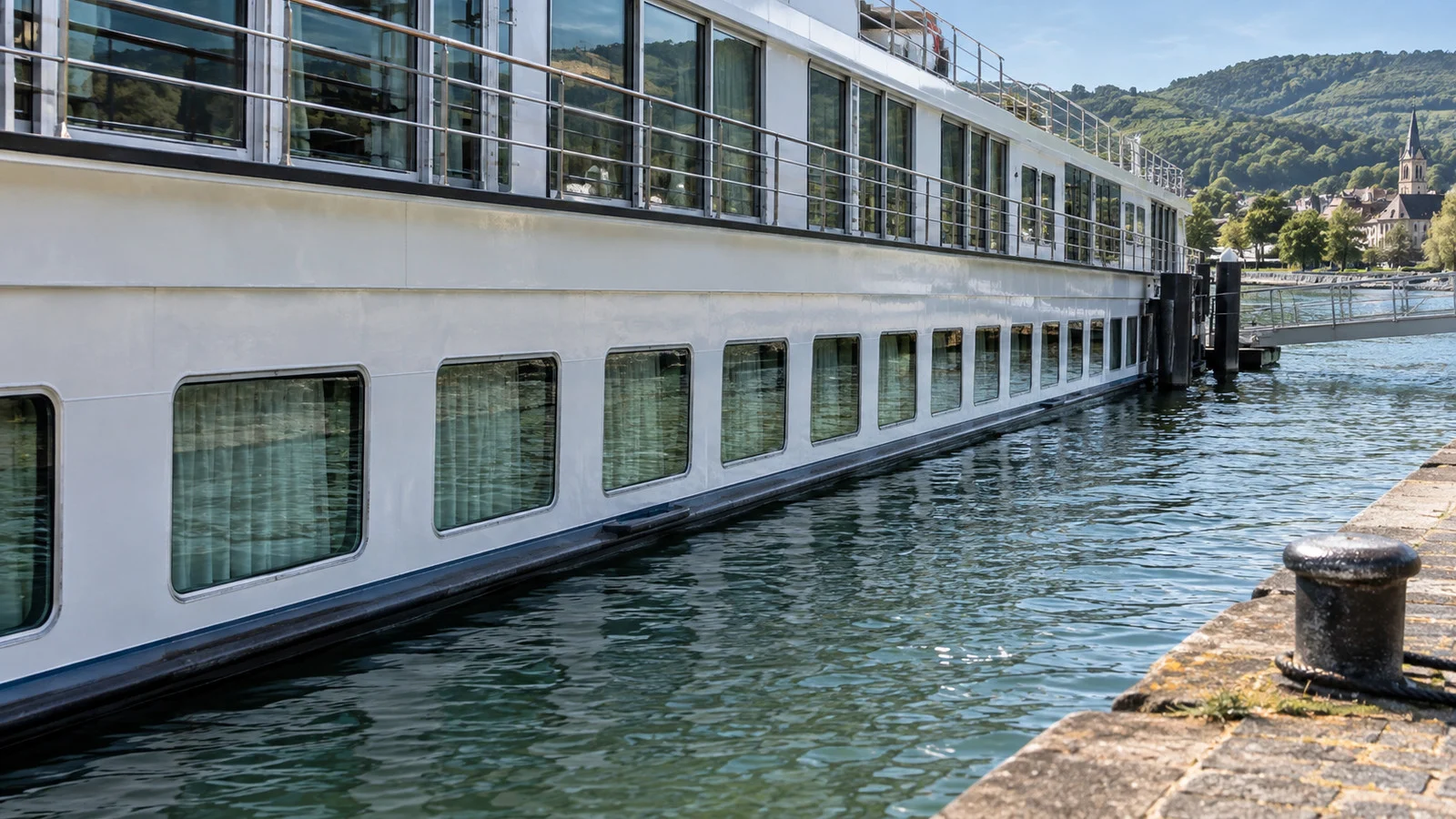 a river cruise ship lower deck exterior at a quiet quay, showing rows of small windows just above the waterline