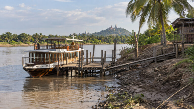 a small Mekong river cruise vessel at a simple wooden rural landing near Kampong Tralach Cambodia, with palm trees and muddy riverbank