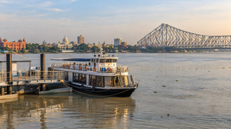 Small river cruise vessel at Kolkata Hooghly River landing with city riverfront and Howrah Bridge in distance, no people