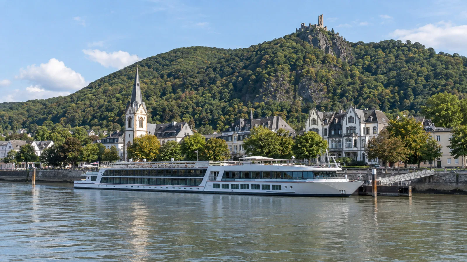 River cruise vessel moored at Konigswinter Germany Rhine waterfront beneath forested Drachenfels hill, no people