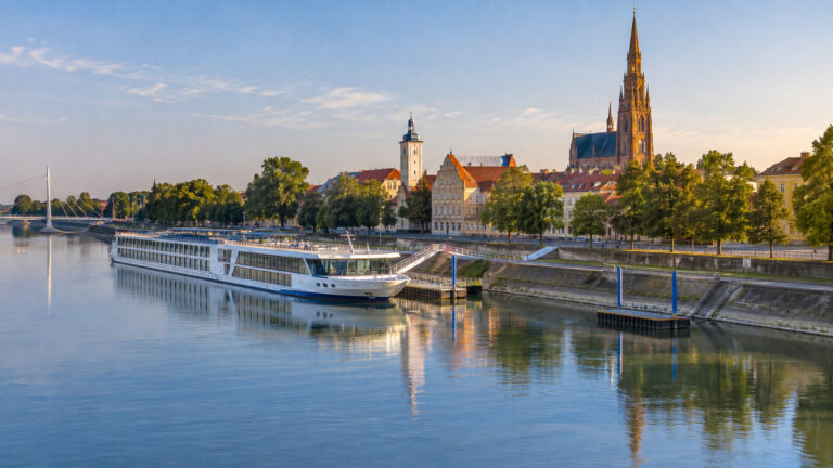 River cruise vessel moored on Drava River at Osijek Croatia with city waterfront and trees, no people