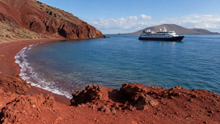 Small Galapagos expedition cruise vessel anchored offshore of Rabida Island red volcanic coast, no people