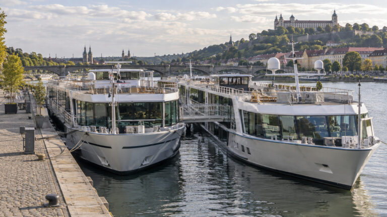 two river cruise ships rafted side by side at a European city quay, one ship's gangway crossing to the other