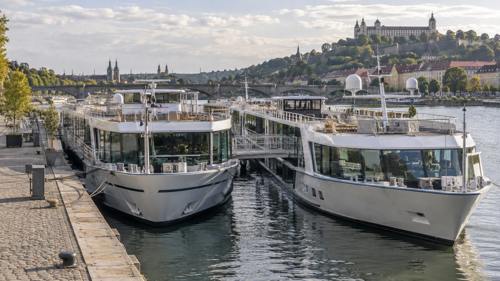 two river cruise ships rafted side by side at a European city quay, one ship's gangway crossing to the other