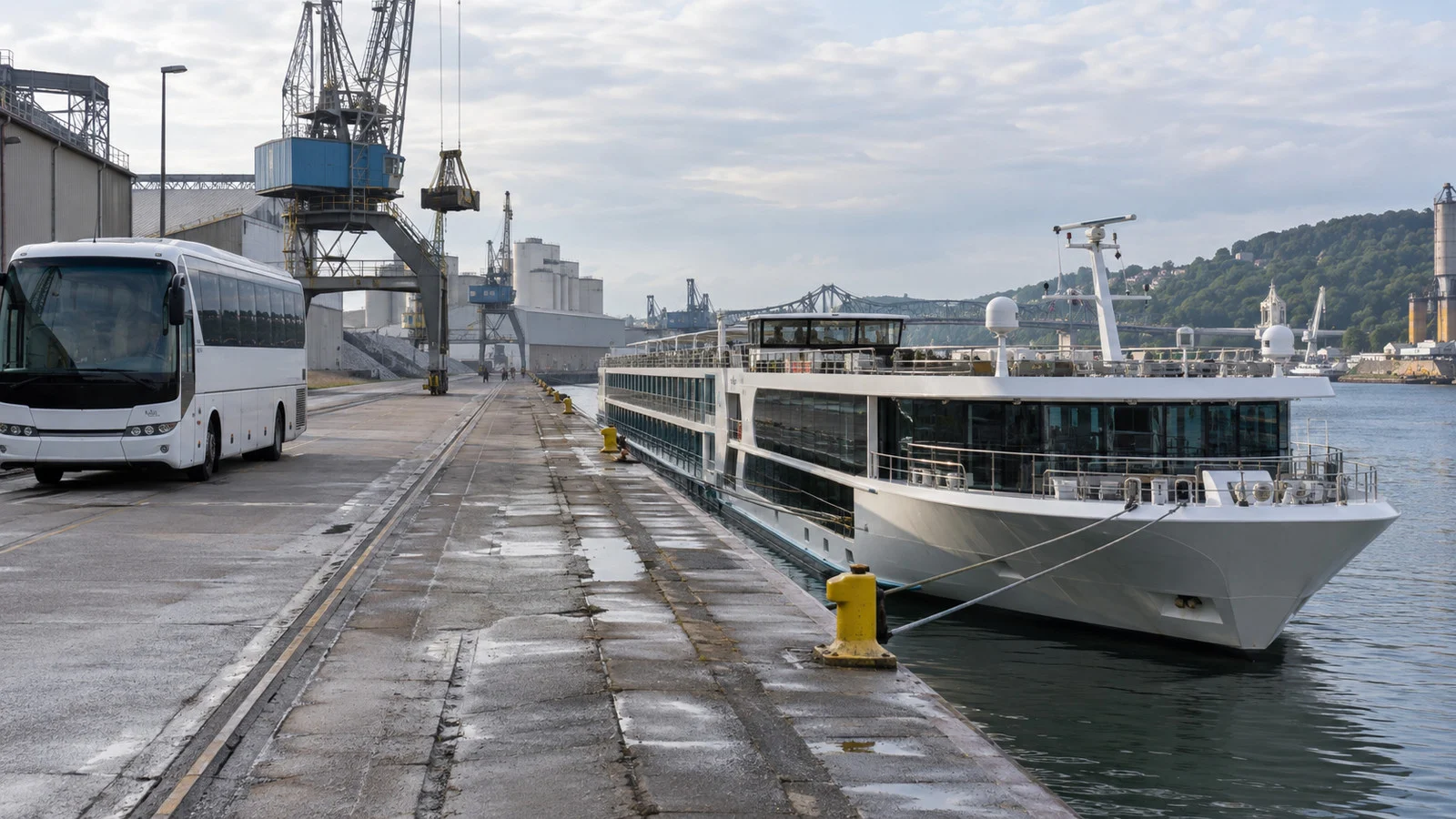a river cruise ship moored at an outer industrial riverside berth with an empty shuttle bus nearby